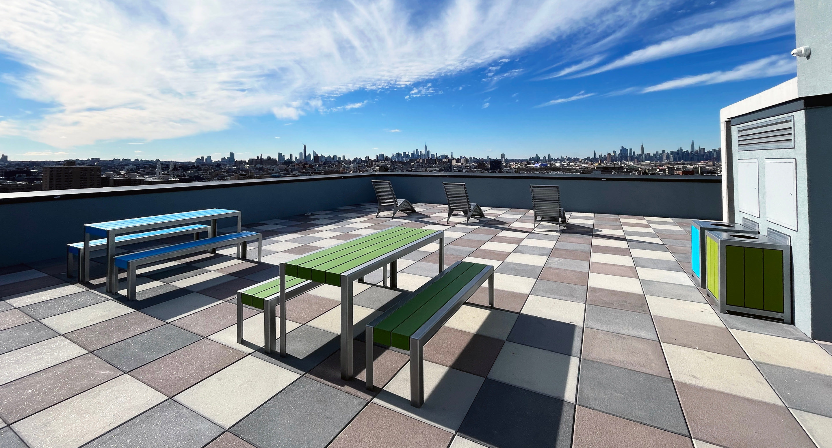 maglin-collections-1050-gallery4 Rooftop patio with checkered tile floor, colorful picnic tables, lounge chairs, and a city skyline in the background under a blue sky with wispy clouds.