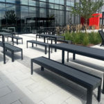 An outdoor patio with black metal tables and benches, modern lounge chairs, greenery, and a glass-front building in the background.