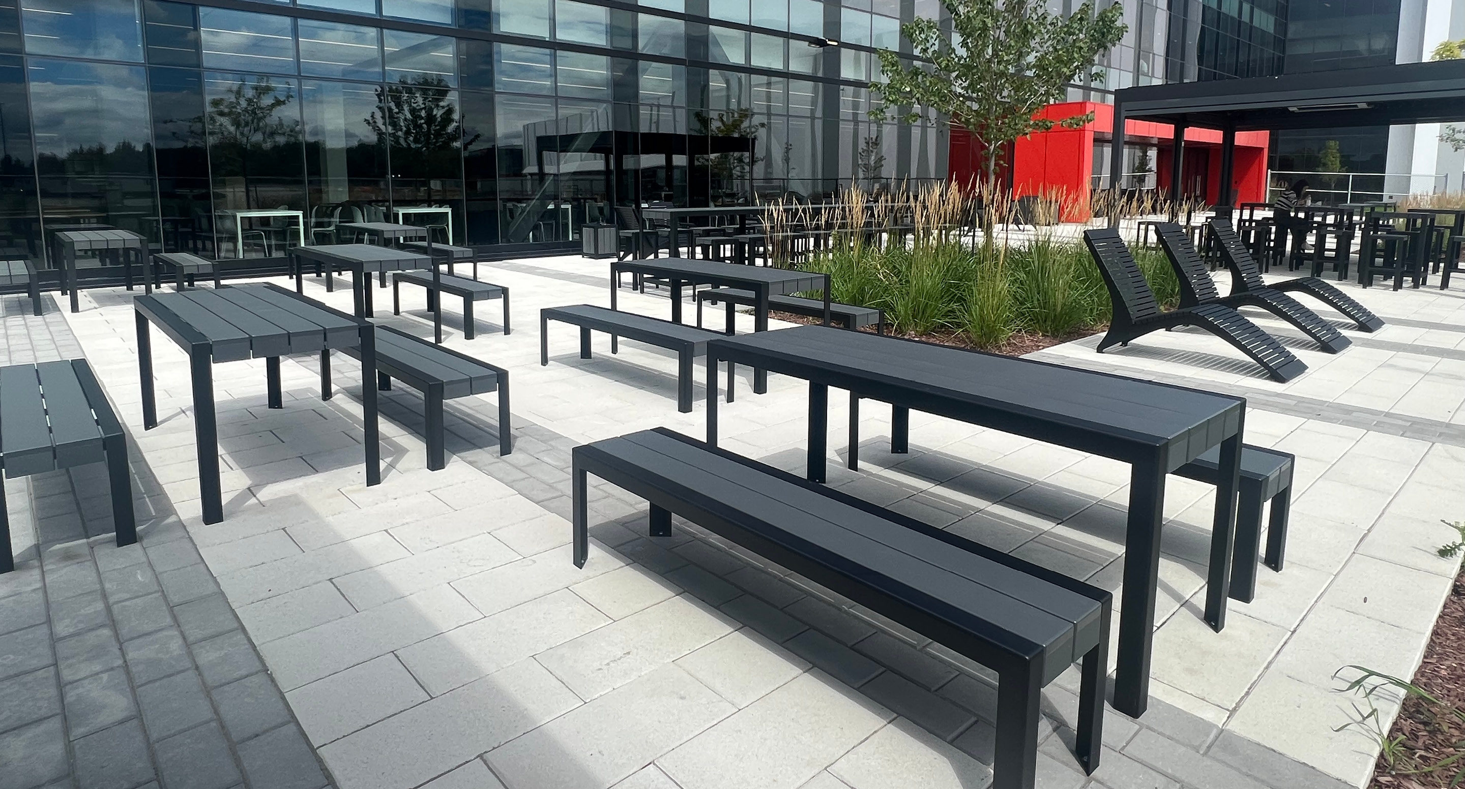 maglin-collections-1050-gallery5 An outdoor patio with black metal tables and benches, modern lounge chairs, greenery, and a glass-front building in the background.