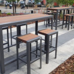 Outdoor patio area with two tall tables and several matching barstools on a paved surface, surrounded by plants, with a street and parked vehicles in the background.