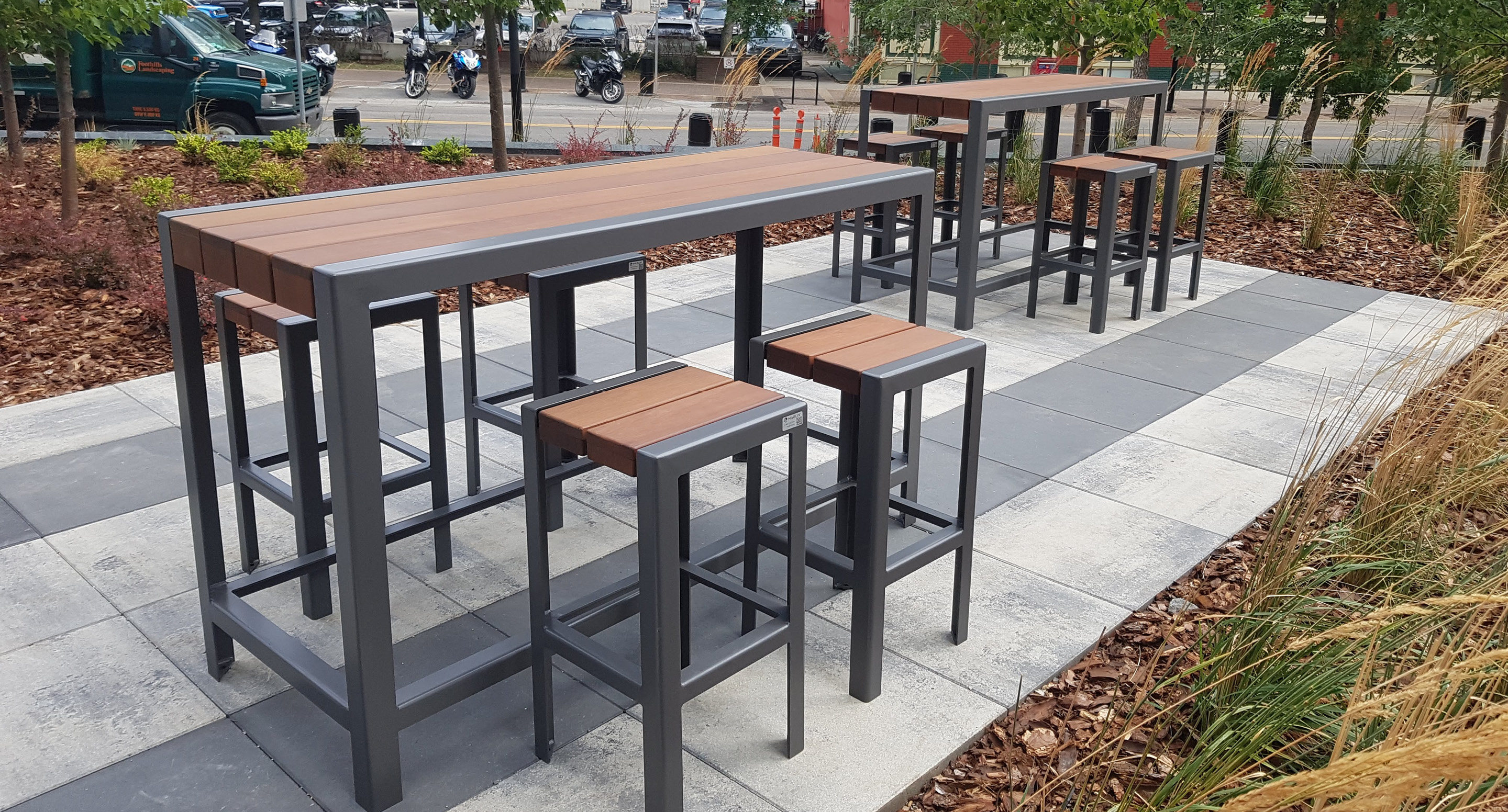 maglin-collections-1050-gallery6 Outdoor patio area with two tall tables and several matching barstools on a paved surface, surrounded by plants, with a street and parked vehicles in the background.