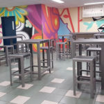 Room with high tables and stools arranged in rows, colorful mural on the walls, tiled floor, and fluorescent ceiling lights.