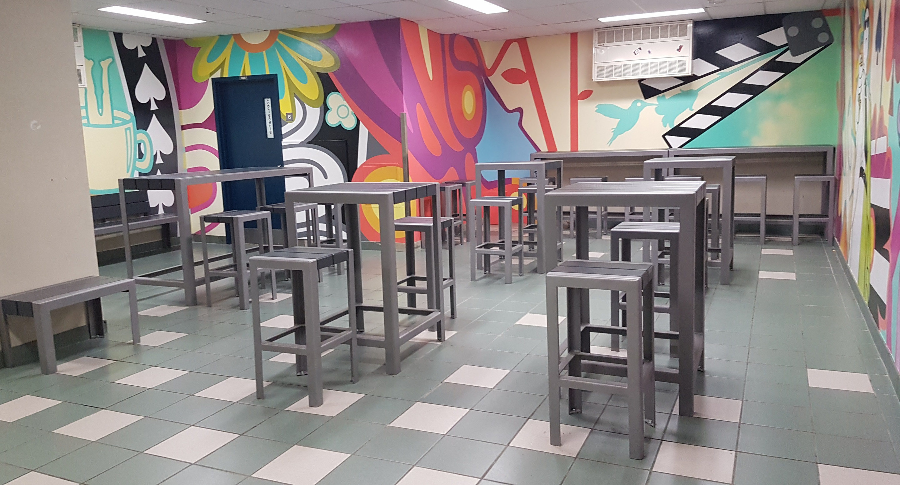 maglin-collections-1050-gallery7 Room with high tables and stools arranged in rows, colorful mural on the walls, tiled floor, and fluorescent ceiling lights.