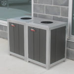 Two rectangular outdoor trash bins with circular openings are placed side by side against a brick wall near a window.