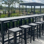 Outdoor patio with black metal tables and stools, shaded pergola, and landscaped greenery on a paved surface under a partly cloudy sky.