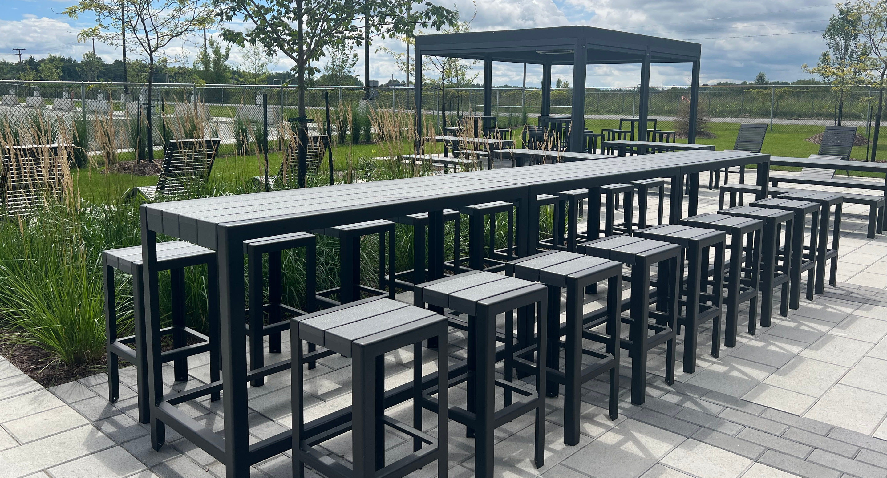 maglin-collections-1050-hero Outdoor patio with black metal tables and stools, shaded pergola, and landscaped greenery on a paved surface under a partly cloudy sky.