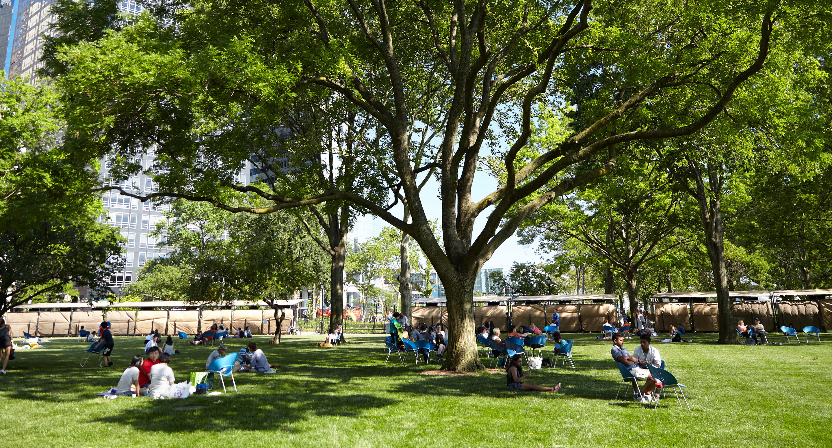 maglin-collections-battery-gallery People sit and relax on chairs under large trees in a green park on a sunny day, with buildings visible in the background.