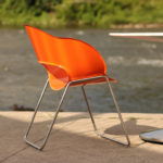 An orange plastic chair with metal legs sits next to a white round table on a concrete surface beside a river.