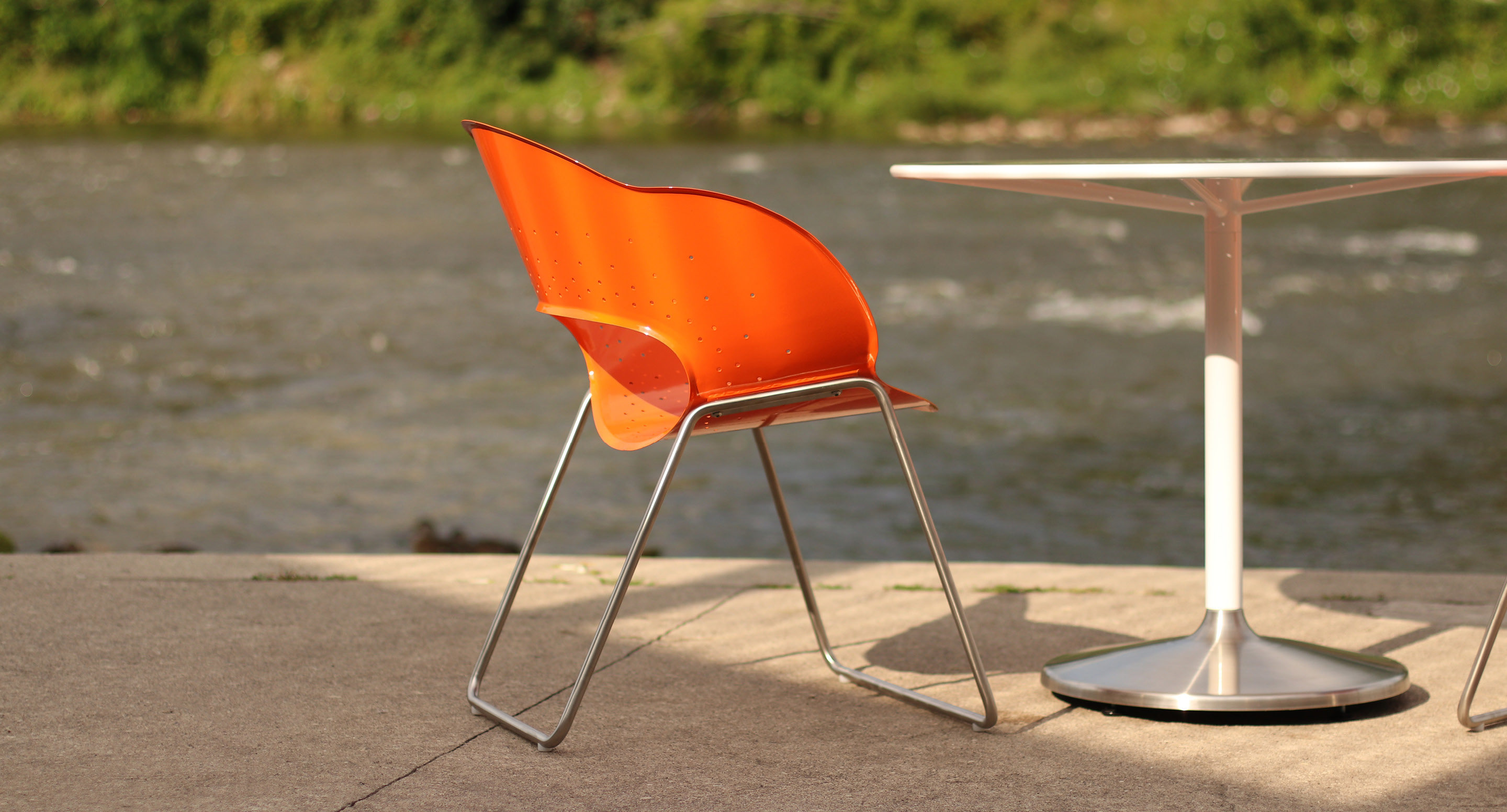 maglin-collections-battery-gallery2 An orange plastic chair with metal legs sits next to a white round table on a concrete surface beside a river.