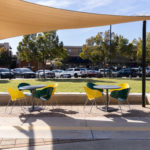 Outdoor seating area with two round tables, yellow and green chairs, and a beige shade canopy, overlooking a grassy field and parked cars.