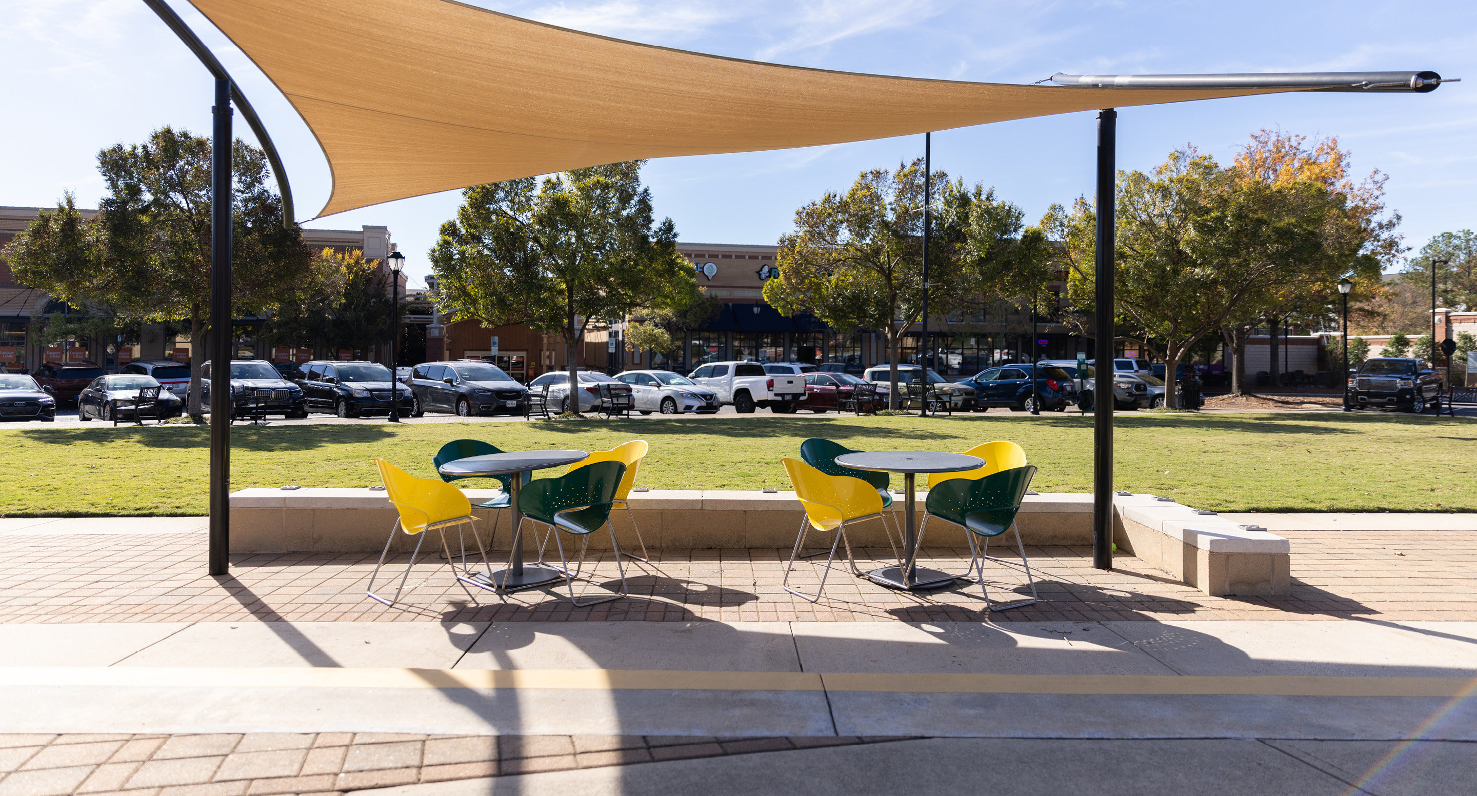 maglin-collections-battery-gallery3 Outdoor seating area with two round tables, yellow and green chairs, and a beige shade canopy, overlooking a grassy field and parked cars.