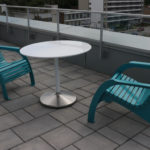Two teal plastic chairs and a round white table are arranged on a tiled outdoor patio with a glass railing overlooking buildings.