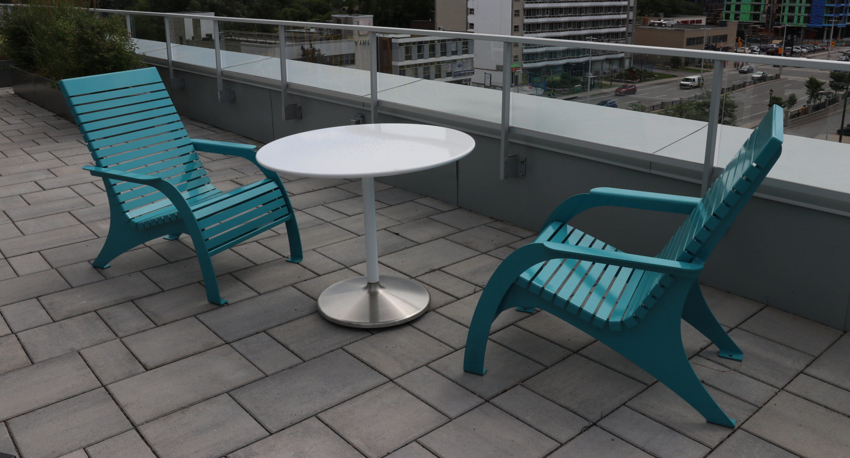 maglin-collections-battery-gallery4 Two teal plastic chairs and a round white table are arranged on a tiled outdoor patio with a glass railing overlooking buildings.