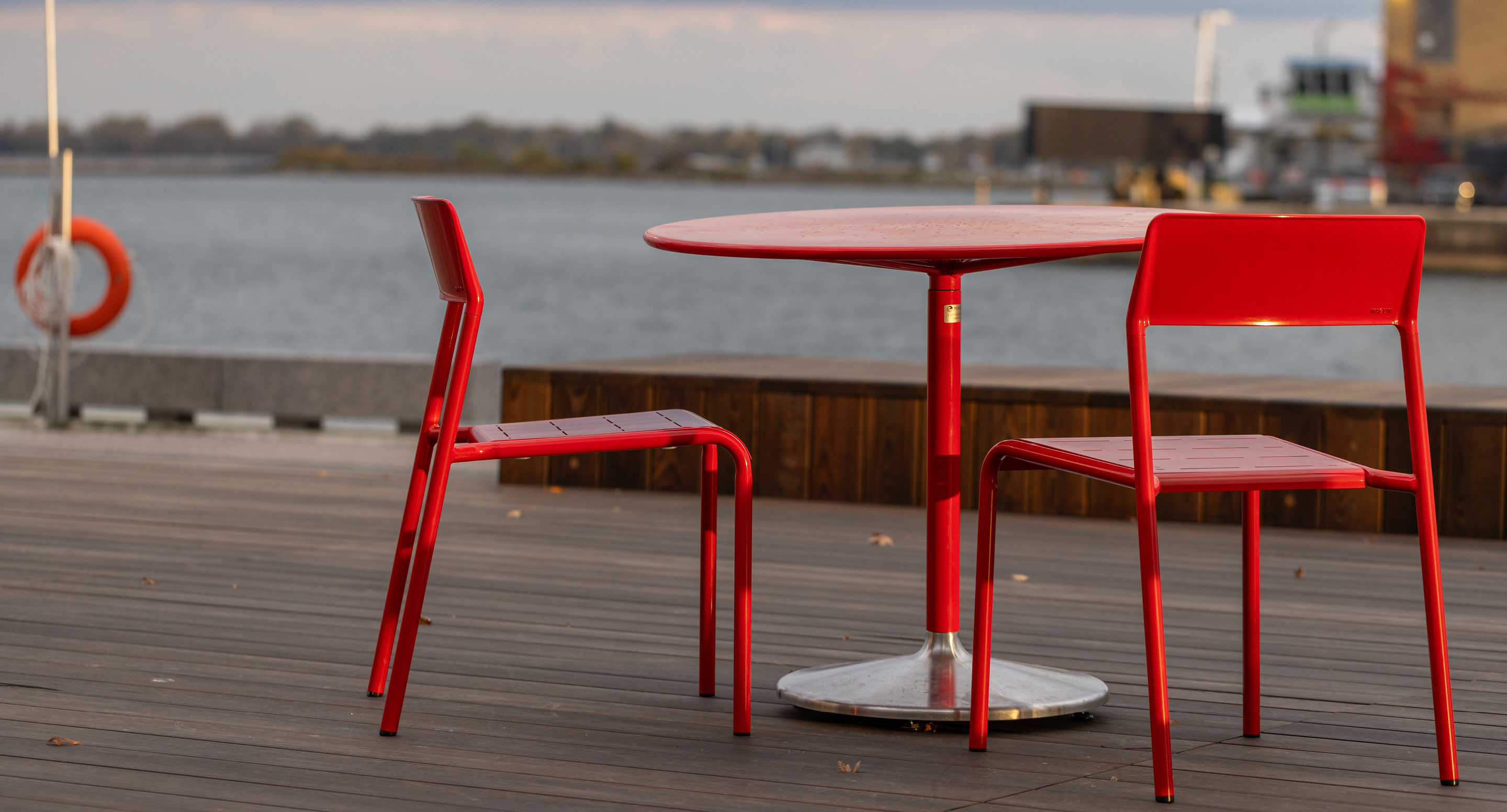 maglin-collections-battery-gallery5 Two empty red chairs and a red round table are placed on a wooden deck overlooking a waterfront with boats and buildings in the background.