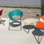 Three colorful modern chairs—red, teal, and orange—are arranged around a small round beige table outdoors on a concrete surface near a beige brick wall.