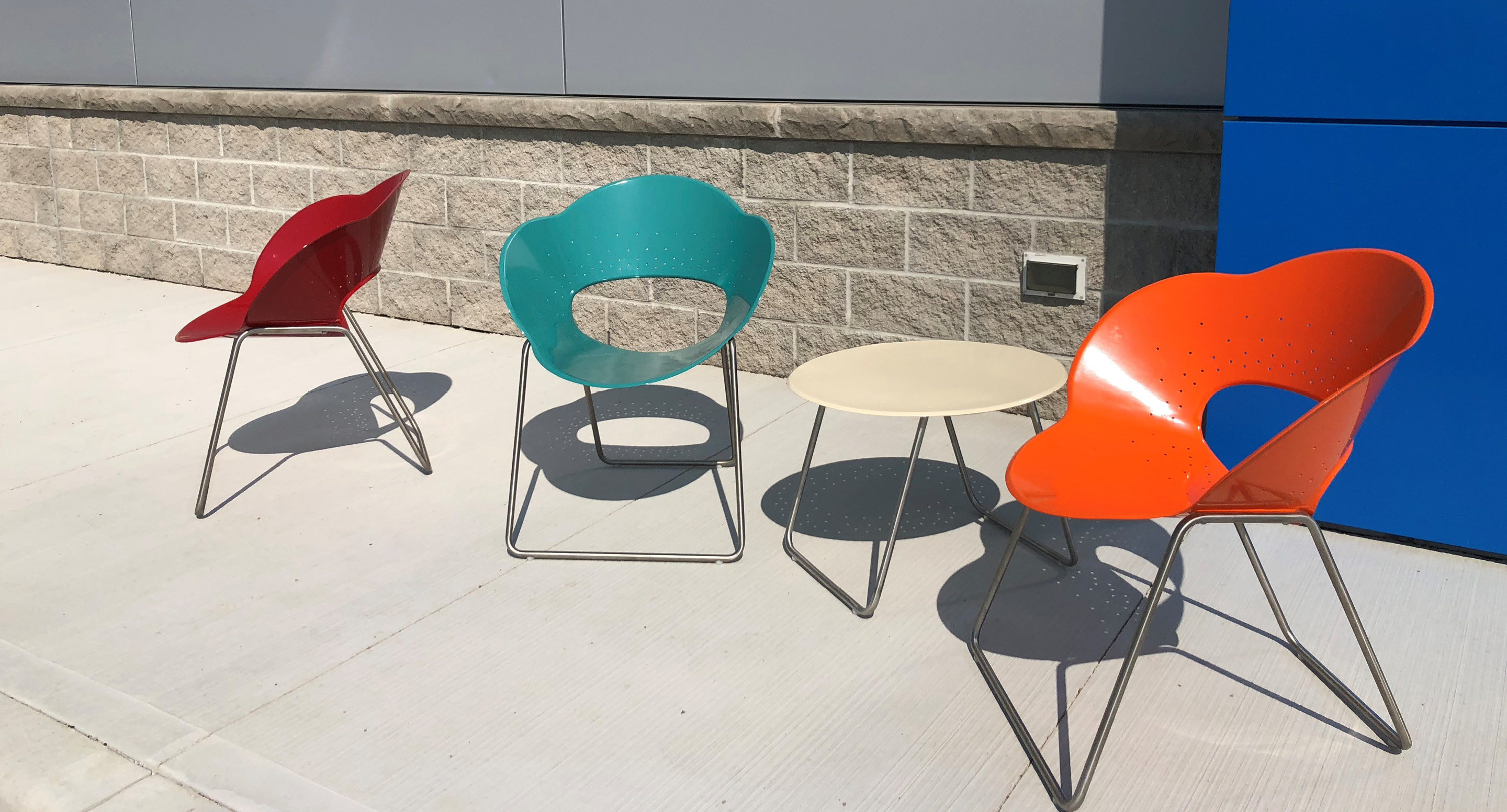 maglin-collections-battery-gallery6 Three colorful modern chairs—red, teal, and orange—are arranged around a small round beige table outdoors on a concrete surface near a beige brick wall.