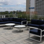 Two navy blue cushioned outdoor sofas and two round white tables are arranged on a rooftop patio with city buildings and greenery in the background.