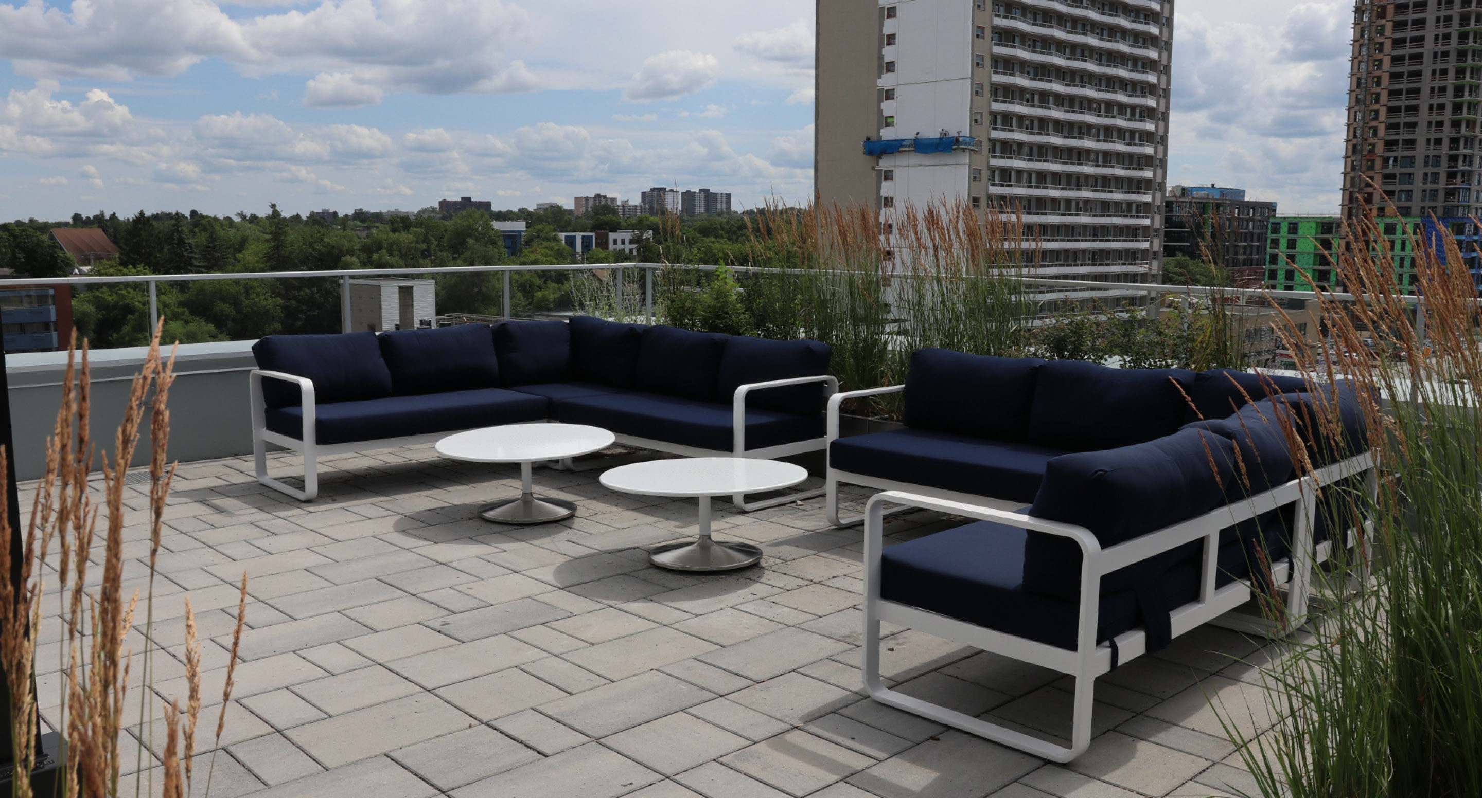 maglin-collections-battery-gallery7 Two navy blue cushioned outdoor sofas and two round white tables are arranged on a rooftop patio with city buildings and greenery in the background.