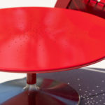 A round red metal table with small holes on its surface sits next to a red chair on a sunny day, casting shadows on a light ground.