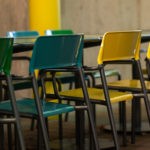 Several rows of green and yellow plastic chairs are arranged around black tables in an outdoor setting with a concrete wall in the background.