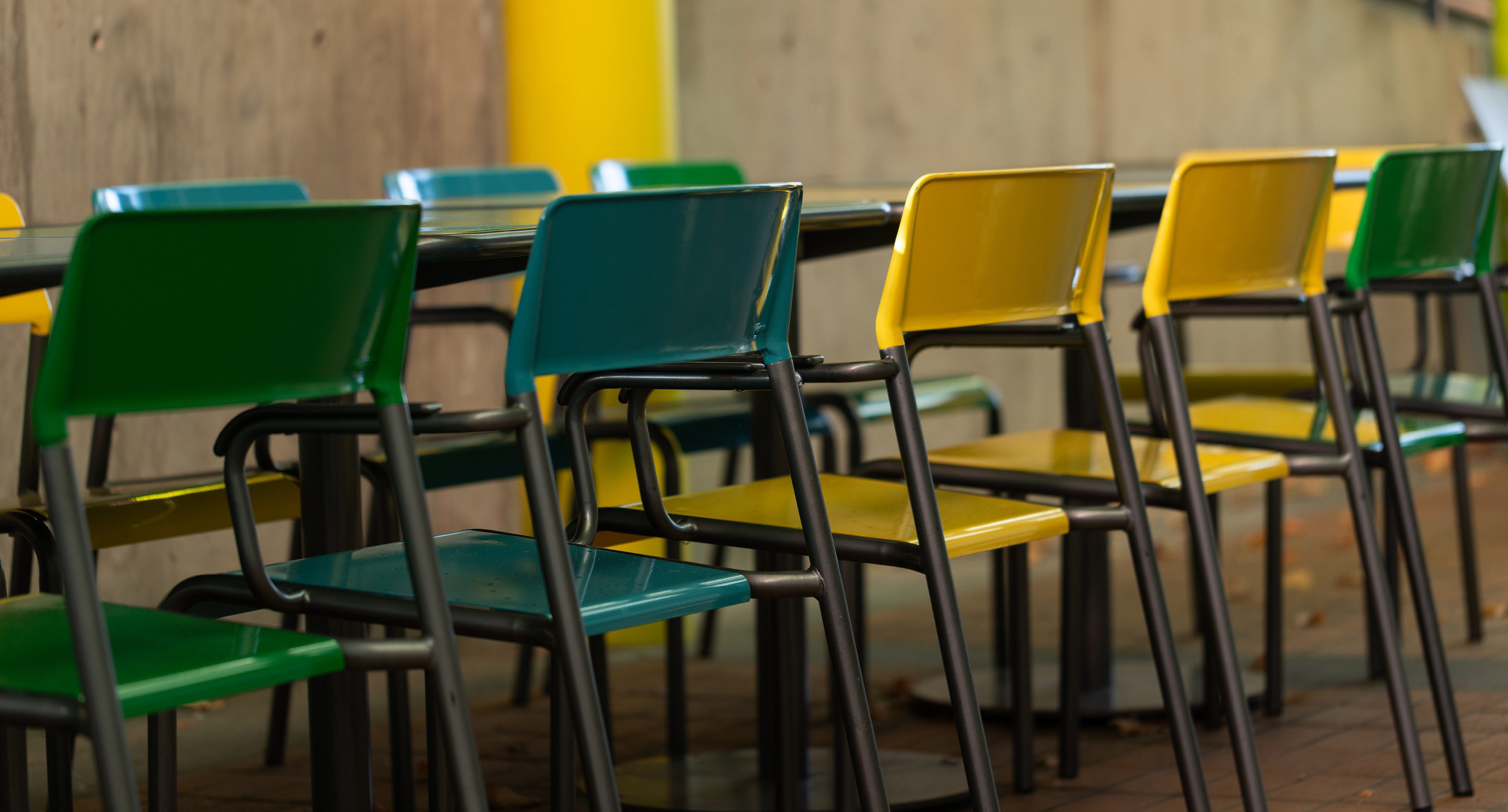 maglin-collections-foro-gallery Several rows of green and yellow plastic chairs are arranged around black tables in an outdoor setting with a concrete wall in the background.