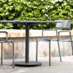 A round black metal table with two matching chairs is set outdoors on a patio, with green bushes and white flowers in the background.