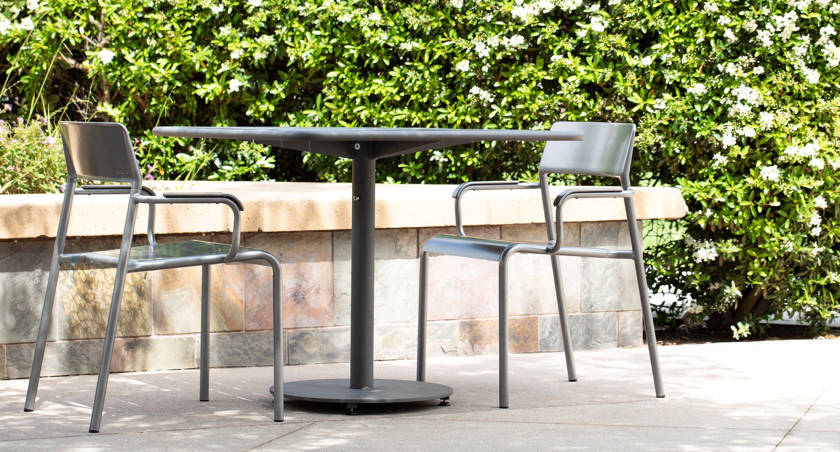 maglin-collections-foro-gallery2 A round black metal table with two matching chairs is set outdoors on a patio, with green bushes and white flowers in the background.