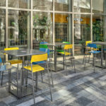 Modern indoor seating area with several metal tables and colorful chairs next to large windows, allowing natural light to fill the space.
