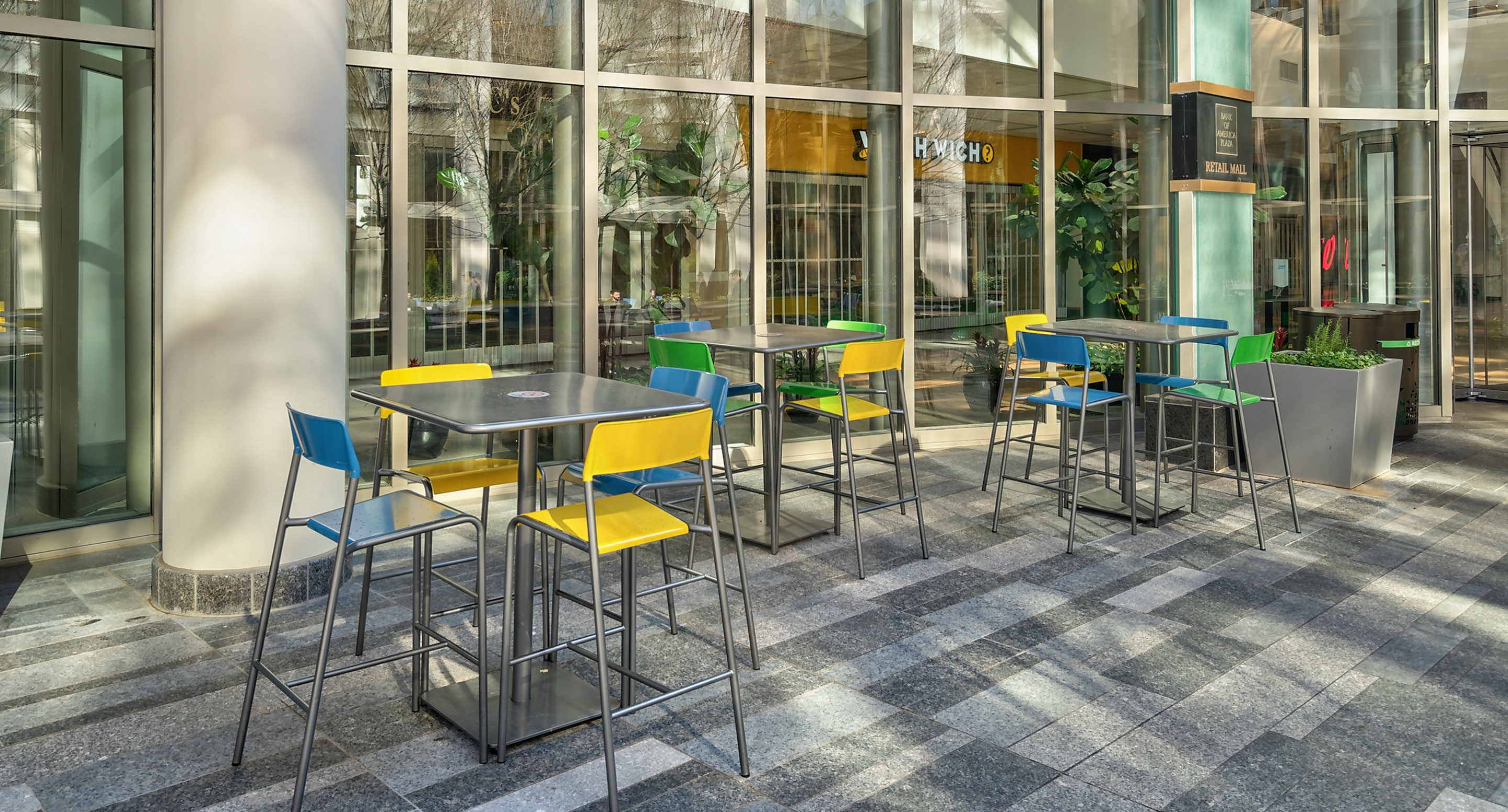 maglin-collections-foro-gallery3 Modern indoor seating area with several metal tables and colorful chairs next to large windows, allowing natural light to fill the space.