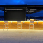 A modern rooftop bar with wooden paneling, high white chairs, and illuminated counter lighting at dusk, overlooking a cityscape.