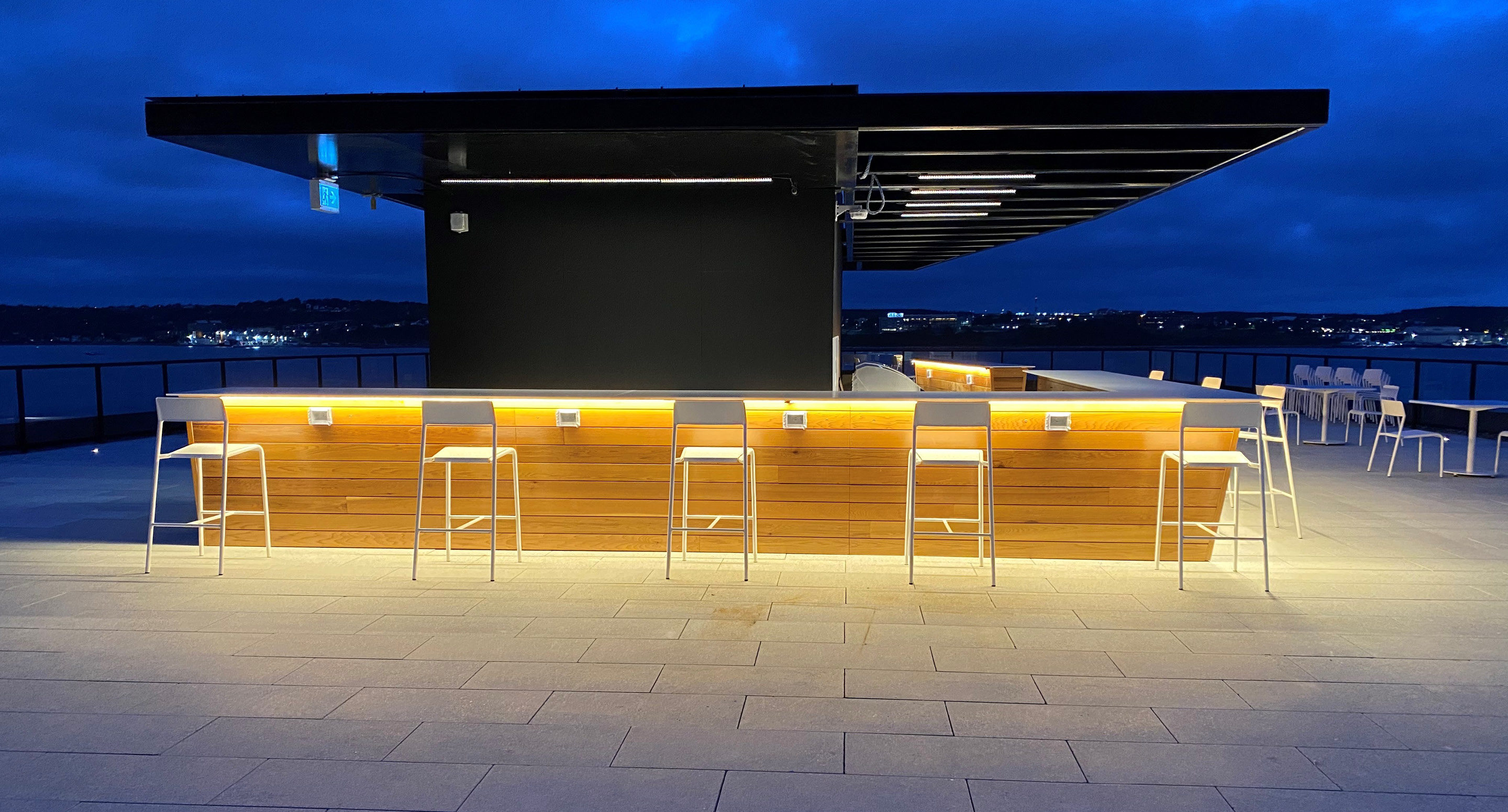 maglin-collections-foro-gallery4 A modern rooftop bar with wooden paneling, high white chairs, and illuminated counter lighting at dusk, overlooking a cityscape.
