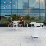 Two people sit at a white table on a modern rooftop terrace with tall buildings and glass windows in the background.