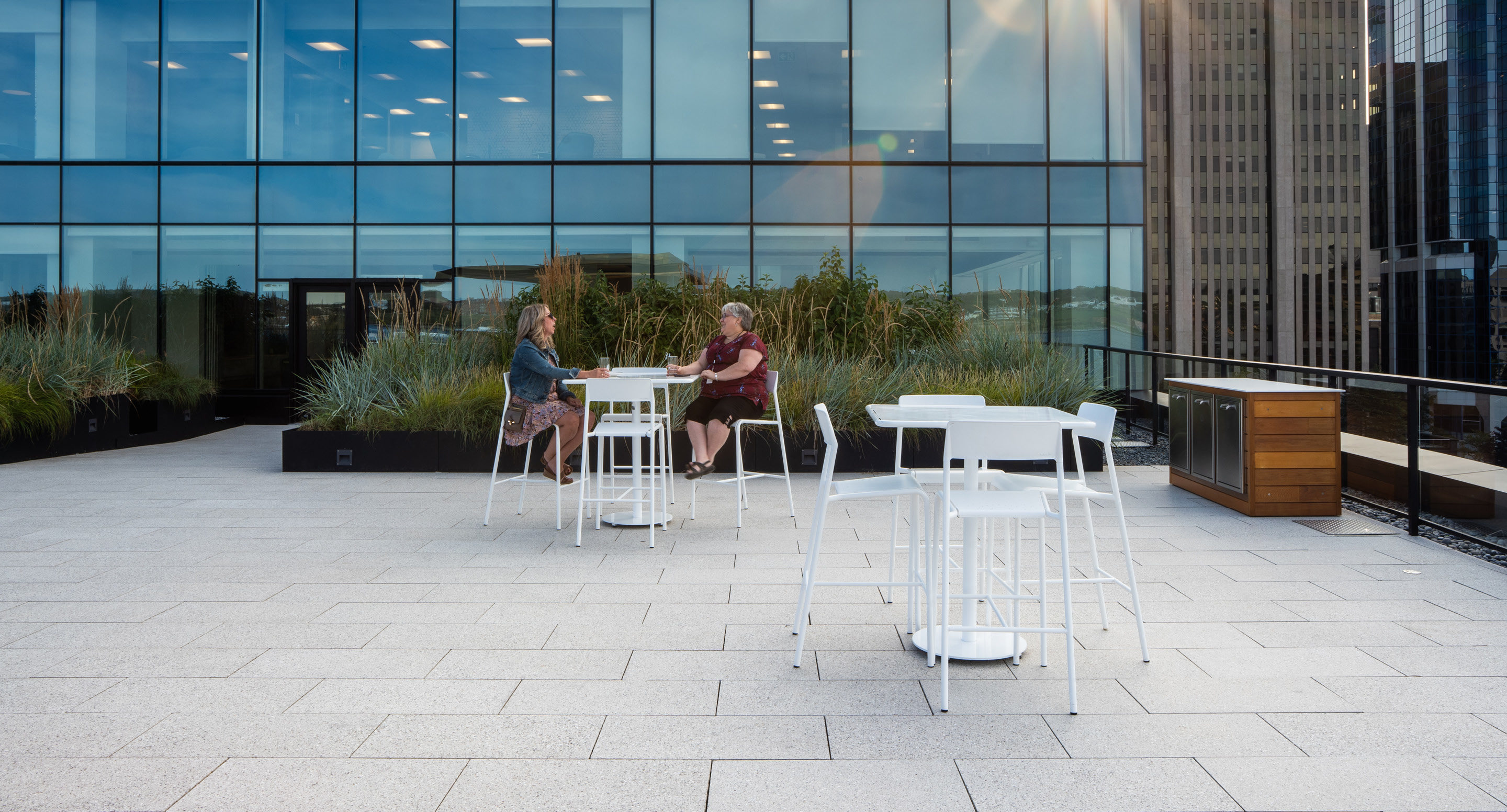 maglin-collections-foro-gallery5 Two people sit at a white table on a modern rooftop terrace with tall buildings and glass windows in the background.