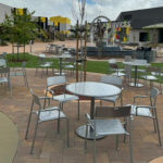 Outdoor plaza with metal tables and chairs on a brick patio, a central fountain, trees, green lawn areas, and modern buildings in the background.