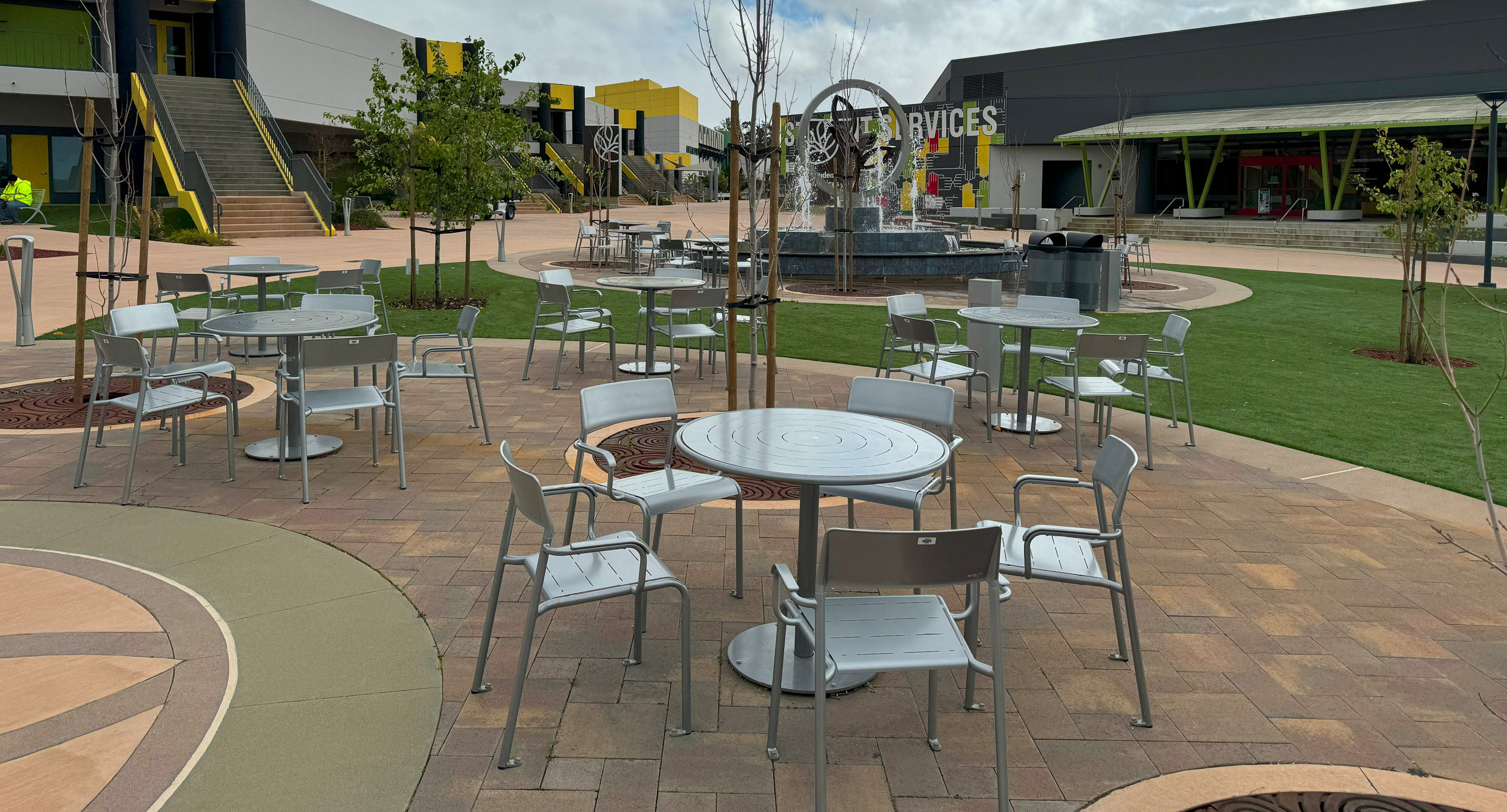maglin-collections-foro-gallery6 Outdoor plaza with metal tables and chairs on a brick patio, a central fountain, trees, green lawn areas, and modern buildings in the background.