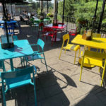 Outdoor patio with colorful tables and chairs in blue, red, and yellow. Menus are placed on some tables. Sunlight casts shadows across the seating area.