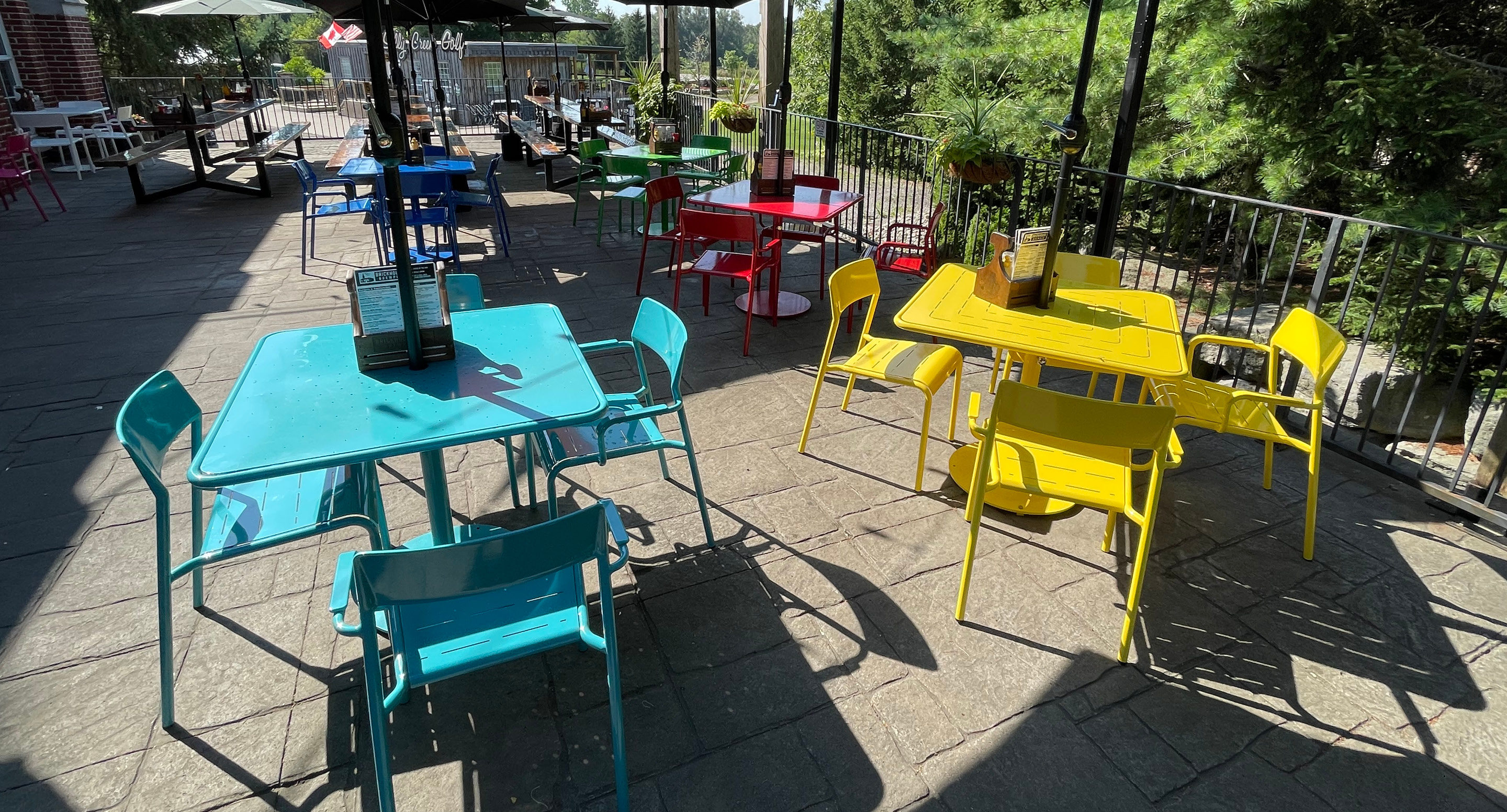 maglin-collections-foro-gallery7 Outdoor patio with colorful tables and chairs in blue, red, and yellow. Menus are placed on some tables. Sunlight casts shadows across the seating area.