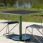 A metal table with a wooden top and two wire chairs are placed on a concrete surface outdoors, with grass and water in the background.