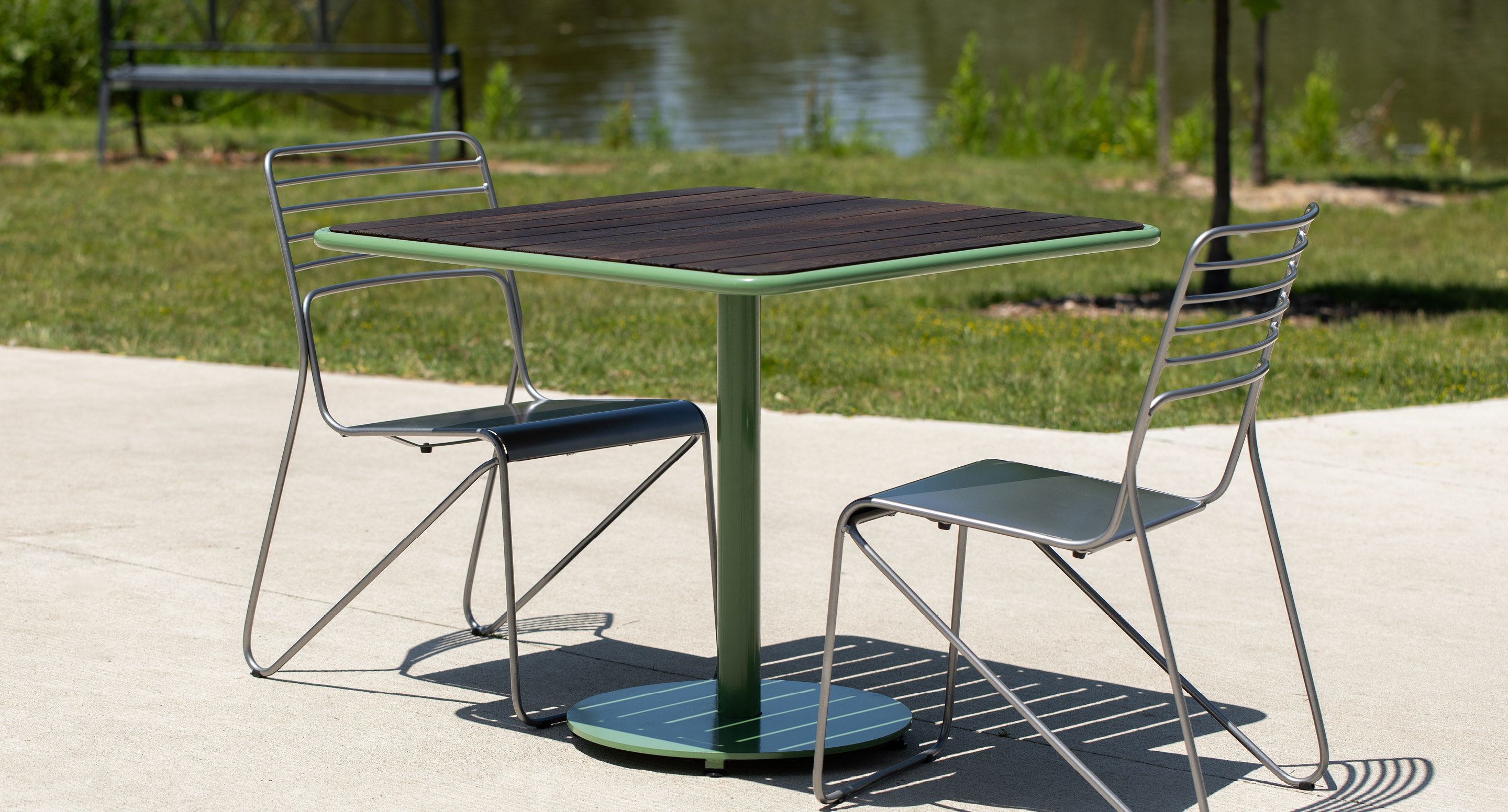 maglin-collections-foro-gallery8 A metal table with a wooden top and two wire chairs are placed on a concrete surface outdoors, with grass and water in the background.