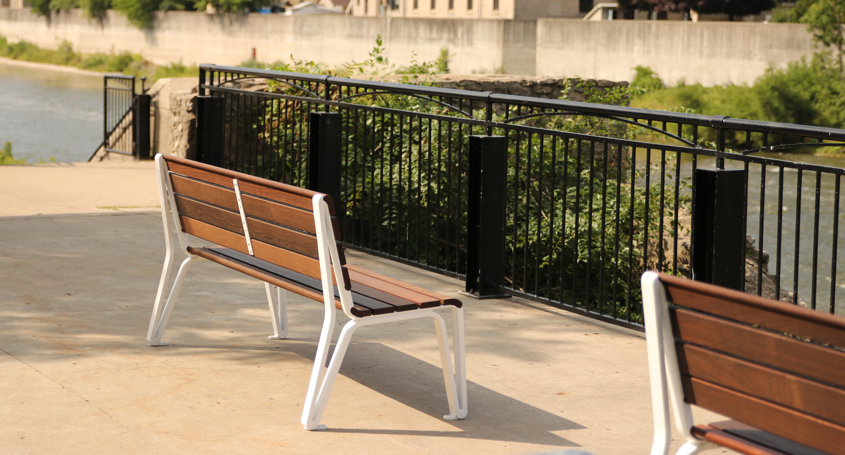 maglin-collections-iconic_gallery Two wooden benches with white metal frames are placed on a paved area near a black metal fence overlooking a river and greenery.