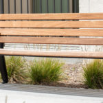 A wooden bench with black metal legs is placed on a concrete sidewalk near some green plants and a building wall.