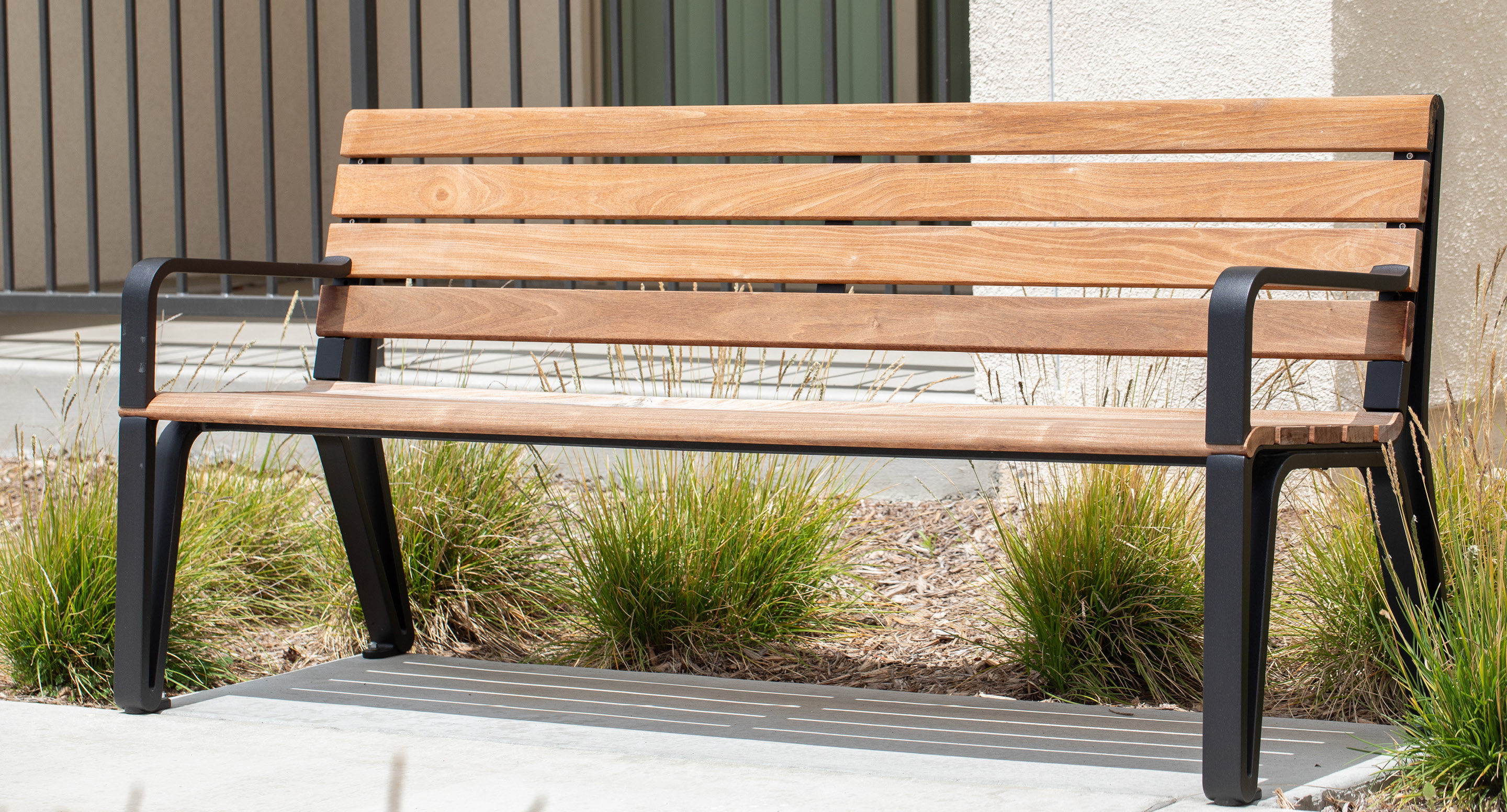 maglin-collections-iconic_gallery3 A wooden bench with black metal legs is placed on a concrete sidewalk near some green plants and a building wall.