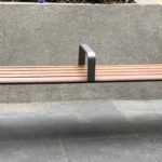 A metal and wooden bench with armrests and a central divider is positioned on a stone pavement in front of a concrete wall.