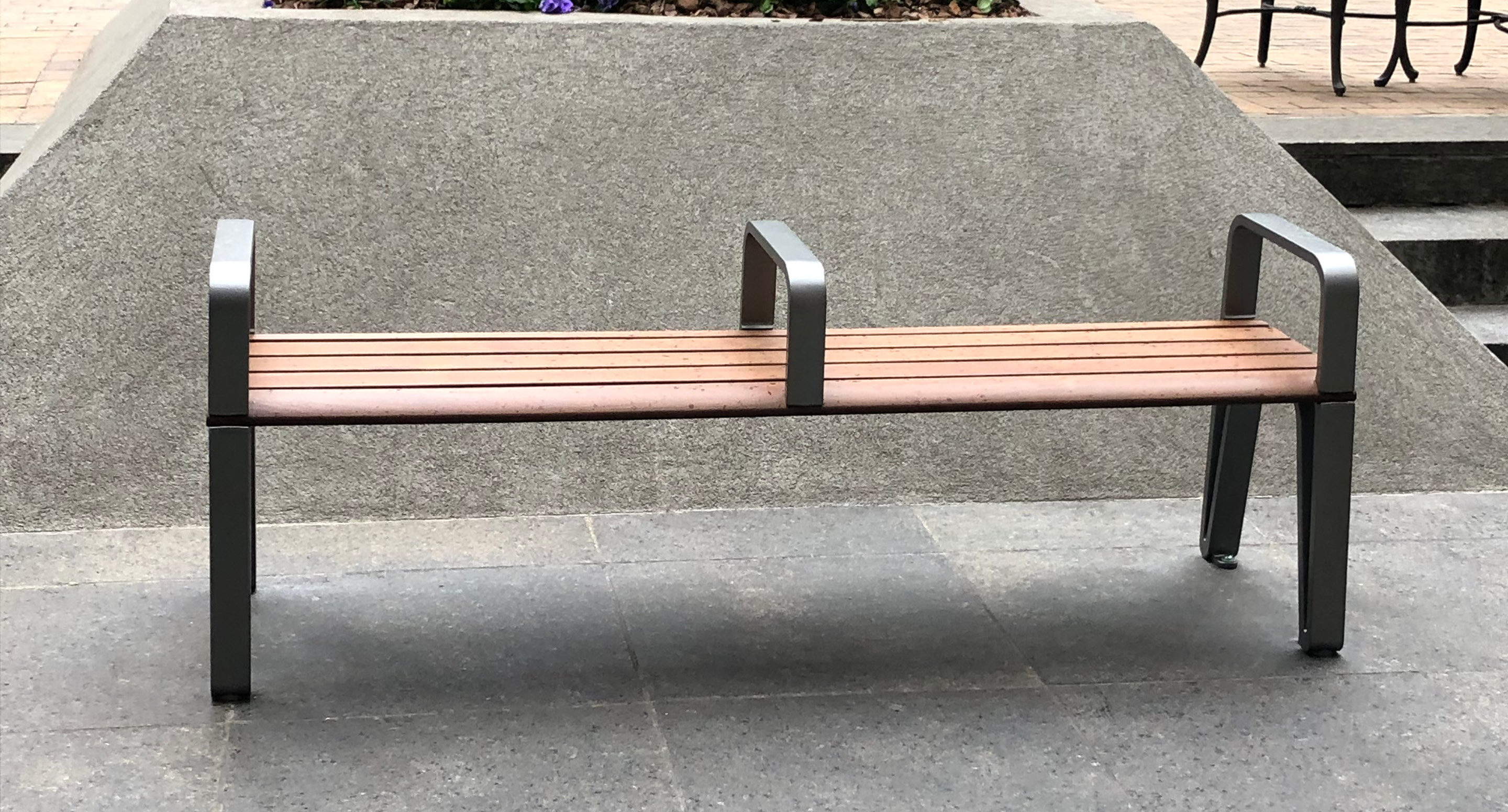 maglin-collections-iconic_gallery4 A metal and wooden bench with armrests and a central divider is positioned on a stone pavement in front of a concrete wall.