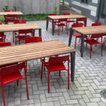 Six rectangular wooden tables with red chairs are arranged on a paved outdoor patio next to a building with striped siding.