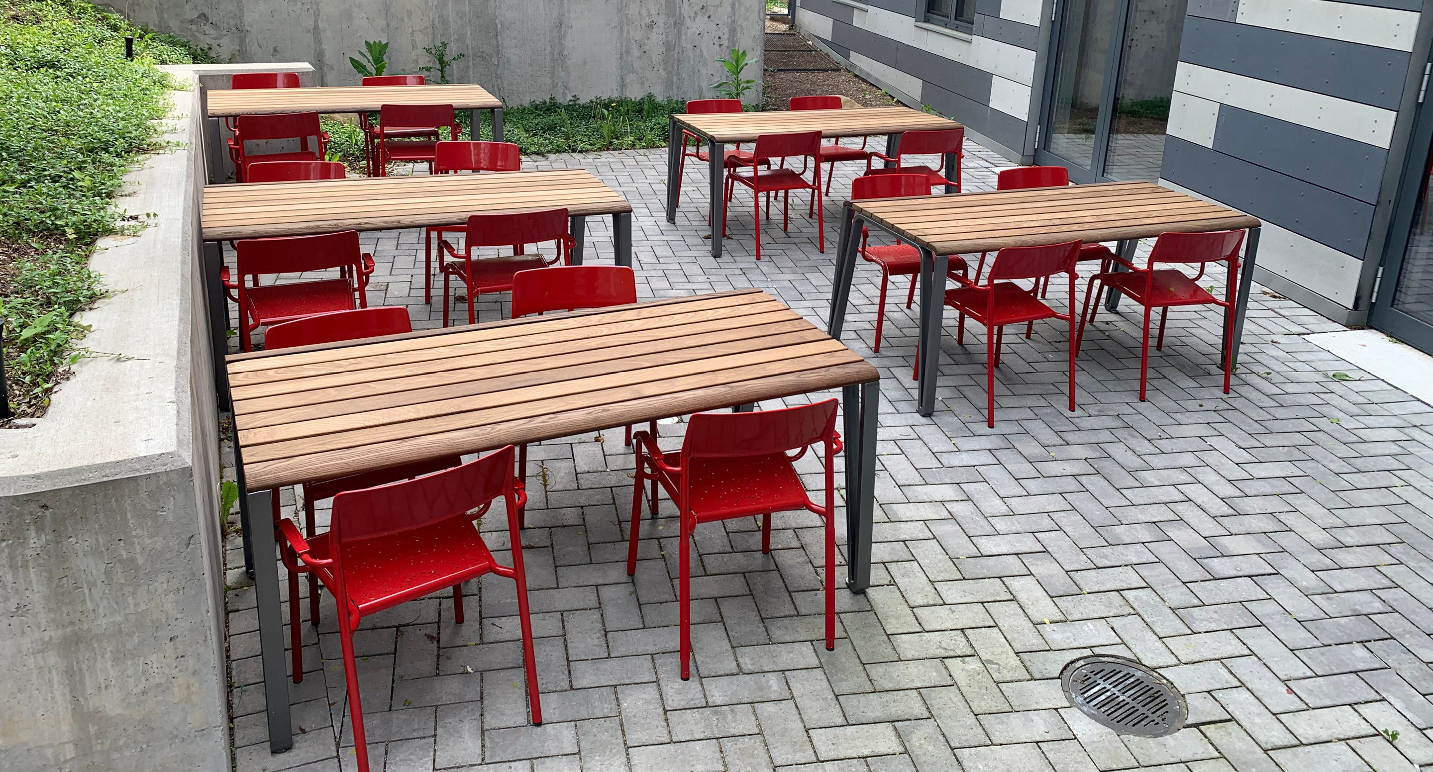 maglin-collections-iconic_gallery5 Six rectangular wooden tables with red chairs are arranged on a paved outdoor patio next to a building with striped siding.