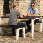 Two men sit facing each other and talking at a wooden picnic table with benches on a cobblestone surface next to a stone building.