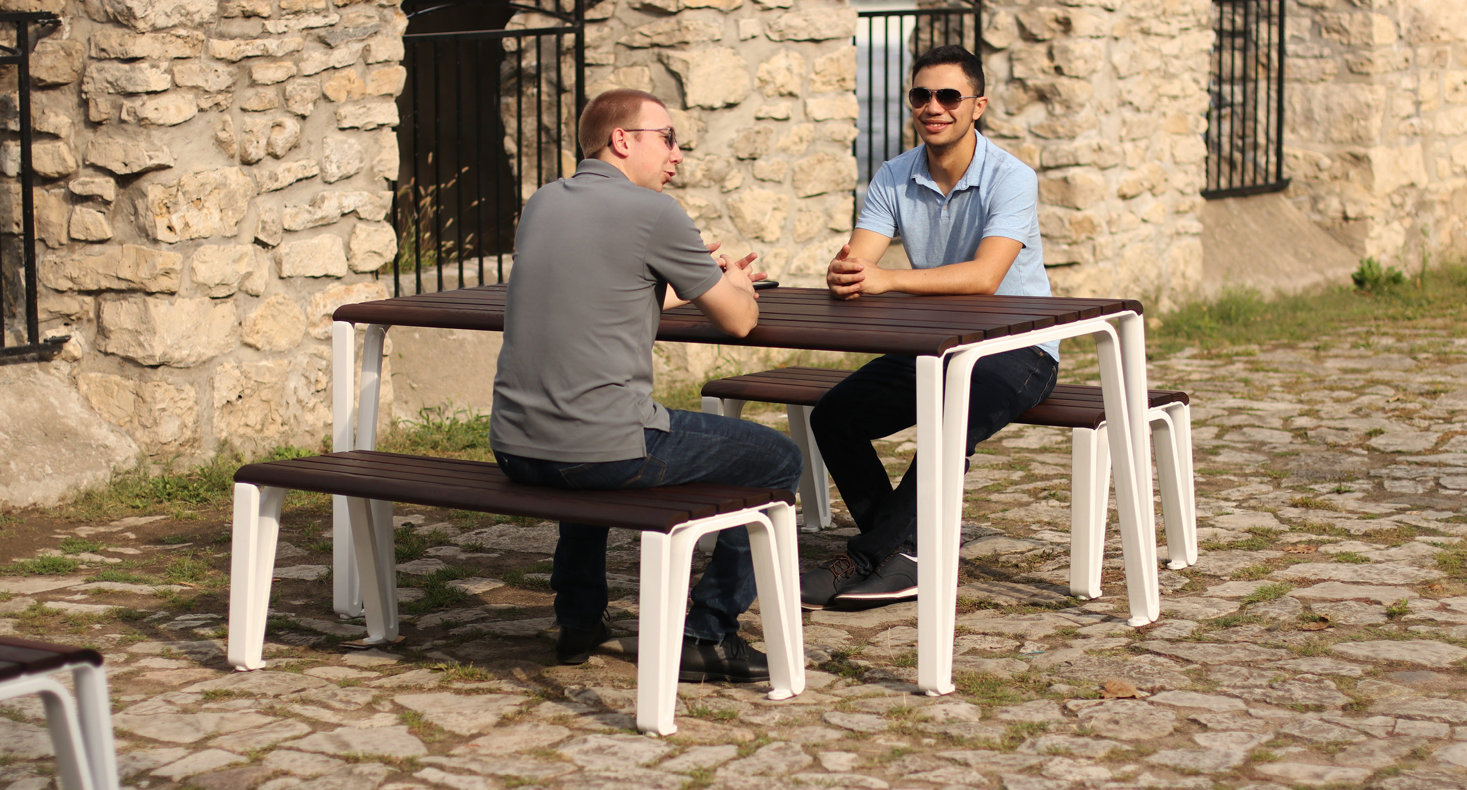 maglin-collections-iconic_gallery6 Two men sit facing each other and talking at a wooden picnic table with benches on a cobblestone surface next to a stone building.