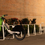 A single white and green bike is parked in a mostly empty bike rack along a sidewalk next to a brick wall with some bushes.