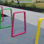 Three colorful metal bike racks—pink, green, and yellow—are installed on a sidewalk in an outdoor park area with grass, shrubs, and a basketball court in the background.